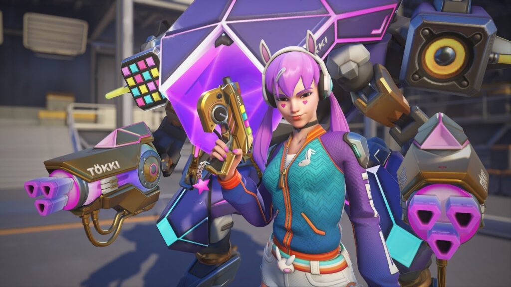 D.Va in her EDM skin from Overwatch 2, featuring vibrant purple hair, a heart-shaped face tattoo, colorful DJ-themed outfit, and a futuristic mech with neon accents and speaker-like elements in the background.